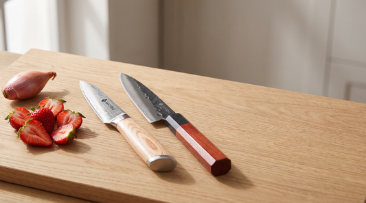 A Japanese-steel petty knife and paring knife laid side by side on a pale oak board with halved strawberries and a shallot